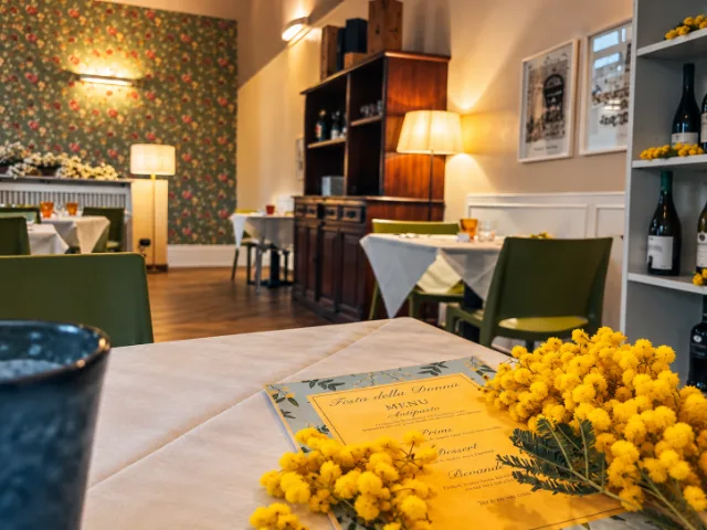 Tavolo del Theorema Ristorante Pizzeria a Pero, con menù dedicato alla Festa della Donna e fiori di mimosa gialli in primo piano, in una sala accogliente con tavoli apparecchiati.