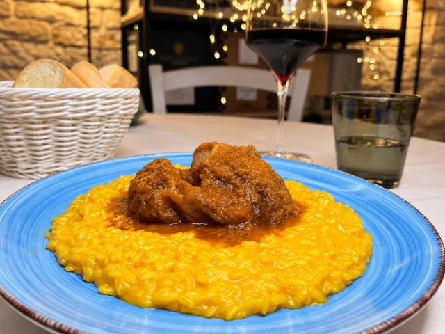 Risotto allo zafferano con carne in umido servito su un piatto blu al Theorema Ristorante Pizzeria a Pero, accompagnato da vino rosso e pane.