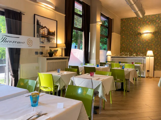 Sala interna del Theorema Ristorante Pizzeria a Pero, con tavoli apparecchiati, sedie verdi, ampie finestre e parete floreale decorativa.