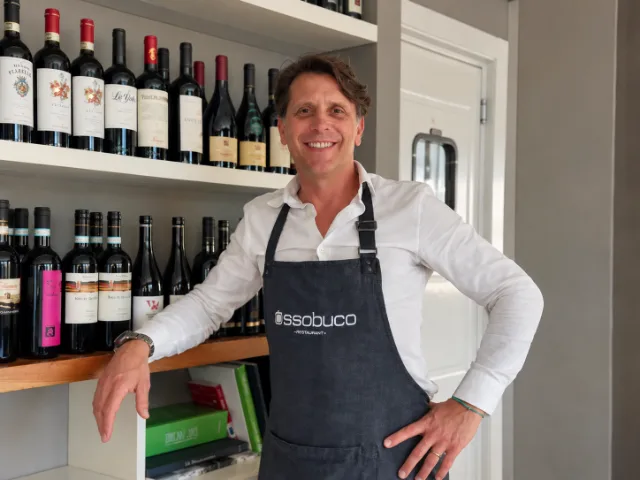 Roberto Monti, fondatore del Ristorante Ossobuco a Varese, sorridente davanti alla selezione di vini del locale, con il grembiule personalizzato Ossobuco.