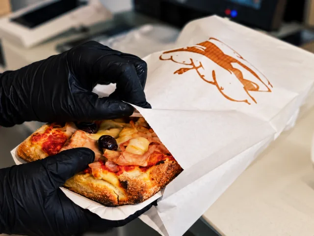 Personale di StàSchiscia Street Food a Milano mentre confeziona una pinsa farcita con prosciutto, formaggio e olive per l’asporto.
