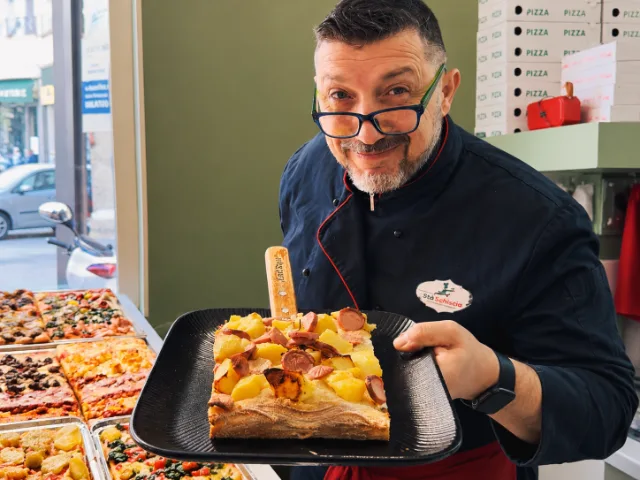 Il titolare di StàSchiscia Street Food a Milano presenta una pizza al trancio farcita con patate e würstel, all’interno del locale.