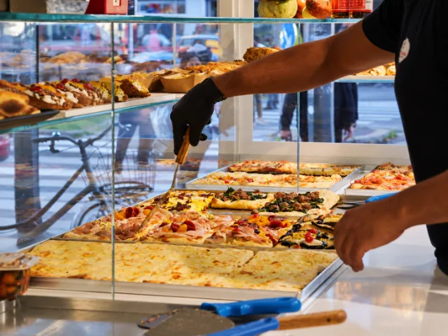 Personale di StàSchiscia Street Food a Milano mentre serve pizze al trancio con diverse farciture dalla vetrina espositiva del locale.