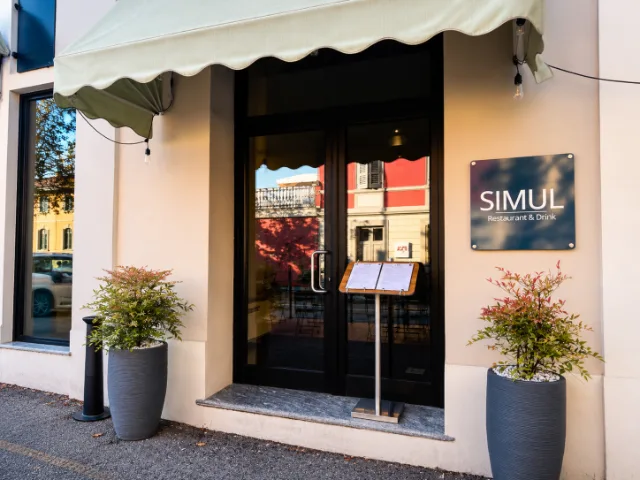 Ingresso di SIMUL Restaurant & Drink a Crema, con insegna del locale, porta a vetri, menù esposto e piante ornamentali ai lati dell'entrata.