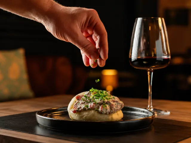 Preparazione di un piatto della cucina contemporanea di SIMUL Restaurant & Drink a Crema, con una mano che aggiunge erba cipollina fresca e un calice di vino rosso accanto.