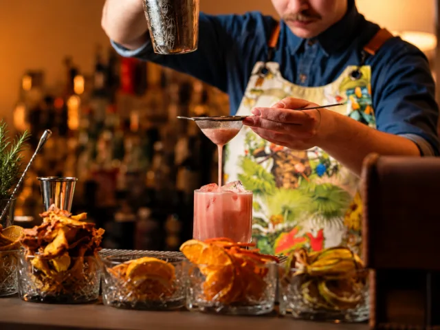 Bartender di SIMUL Restaurant & Drink a Crema mentre prepara un cocktail rosa, versandolo attraverso un colino nel bicchiere con ghiaccio, tra ingredienti e decorazioni da cocktail.