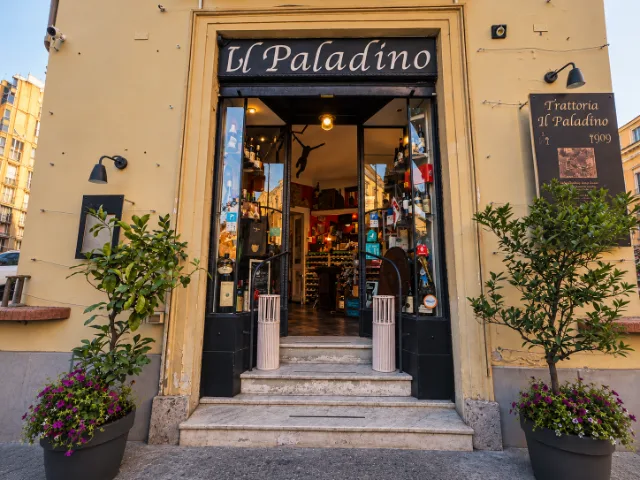 Ingresso della Trattoria Il Paladino ad Alessandria, con insegna storica, gradini in marmo, piante ornamentali e vetrine che lasciano intravedere gli ambienti interni del ristorante.