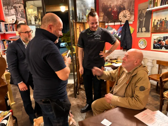 Momento di convivialità all'interno della Trattoria Il Paladino ad Alessandria, con personale e ospiti che conversano in una sala caratterizzata da pareti rosse, fotografie storiche e arredi tradizionali.