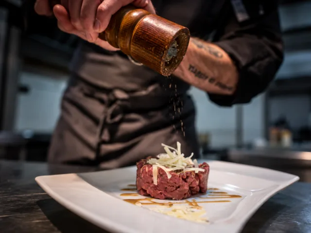 Preparazione di una tartare di carne alla Trattoria Il Paladino di Alessandria, rifinita con scaglie di formaggio e pepe macinato al momento, a testimonianza della cura dedicata a ogni piatto.
