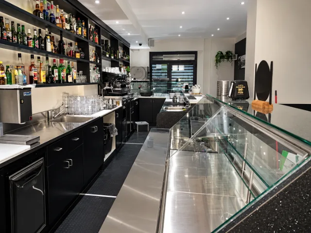 Bar profesional cu vitrină de prezentare din sticlă, echipamente din oțel inoxidabil, espressor și rafturi pentru băuturi.