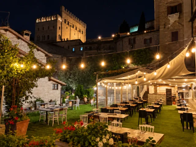 Giardino panoramico del Ristorante Duca Federico da Montefeltro a Gubbio, con tavoli all'aperto, luci calde e vista sul Palazzo dei Consoli illuminato di sera.