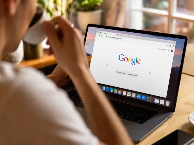 Persona che utilizza un computer portatile per effettuare una ricerca su Google, seduta a una scrivania con una tazza di caffè.