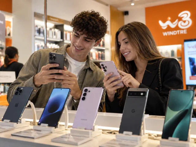 Due clienti confrontano diversi modelli di smartphone Apple e Samsung all'interno di un punto vendita WINDTRE, valutandone caratteristiche e funzionalità.