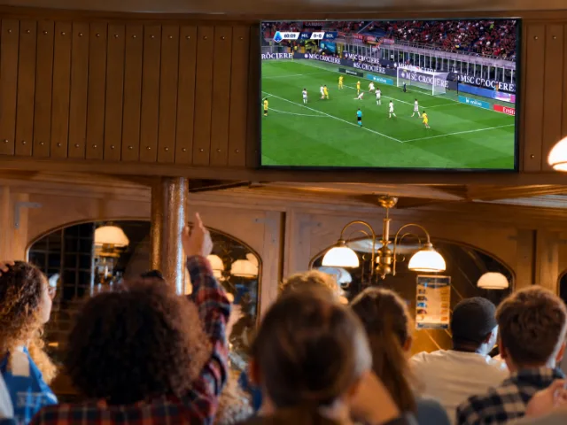Kunden verfolgen ein Serie-A-Fußballspiel, das auf einem großen Fernseher in einem Lokal mit Holzmöbeln übertragen wird.