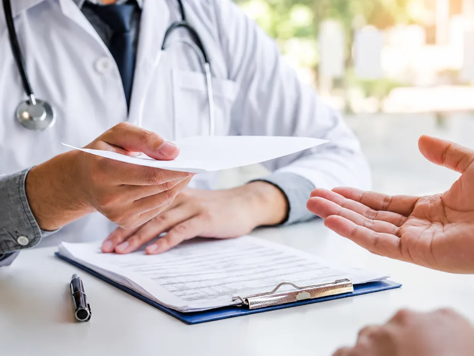 Medico con camice e stetoscopio durante un consulto, mentre consegna un documento a un paziente seduto davanti alla scrivania.